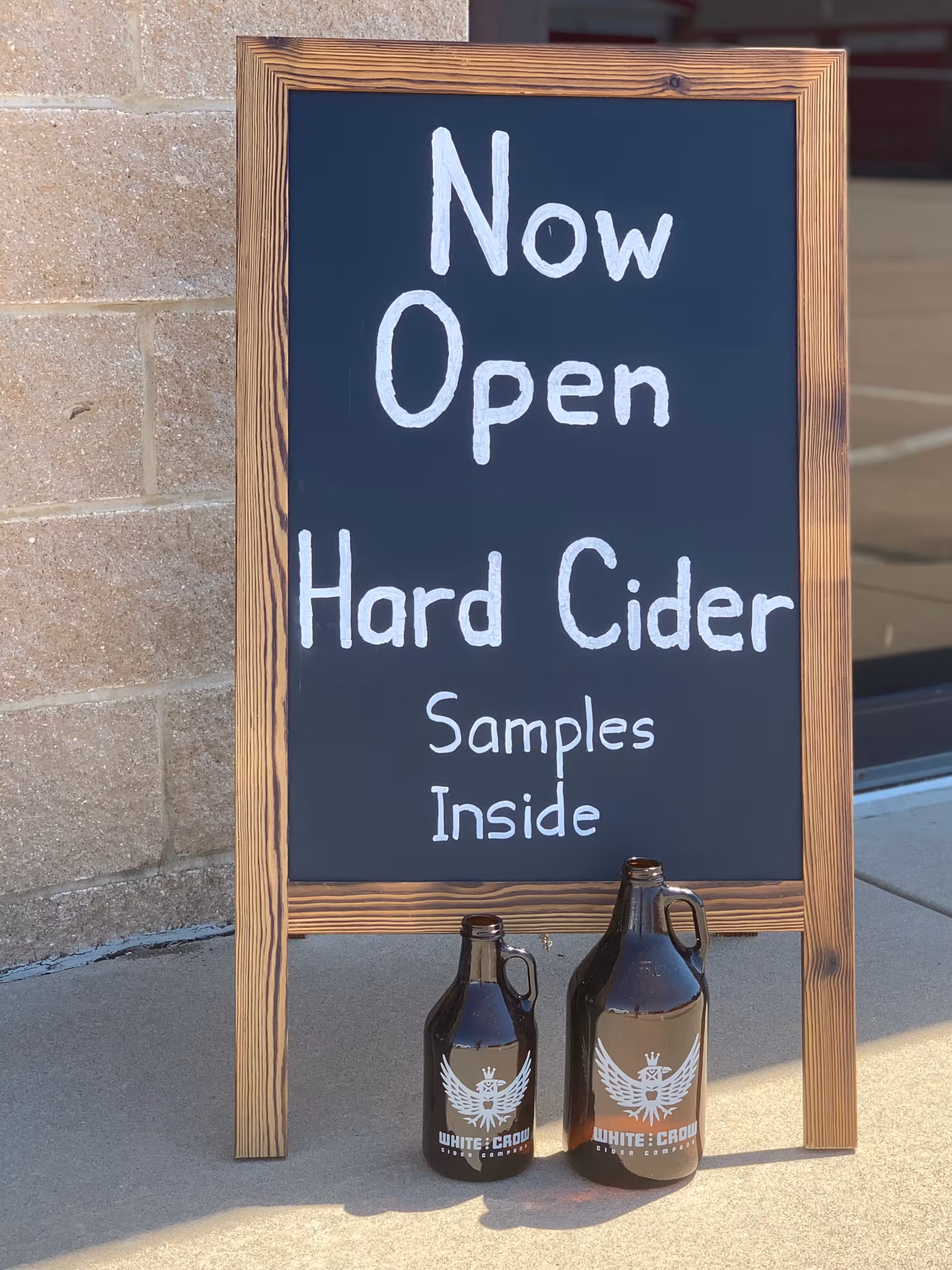 Wooden chalkboard sign reading ‘Now open – hard cider samples inside.’